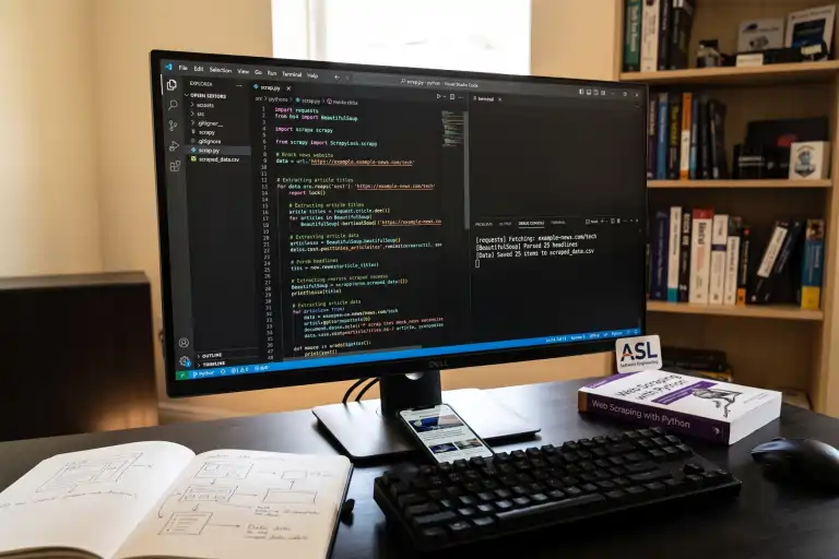 Fotografia do ambiente de trabalho de um programador. O monitor central exibe um script de Web Scraping em Python no editor VS Code, utilizando as bibliotecas Requests e BeautifulSoup. Sobre a mesa escura estão um teclado mecânico, um smartphone, um caderno aberto com rascunhos de interface, um livro impresso sobre Web Scraping com Python e um pequeno display com a logo da ASL Software Engineering.